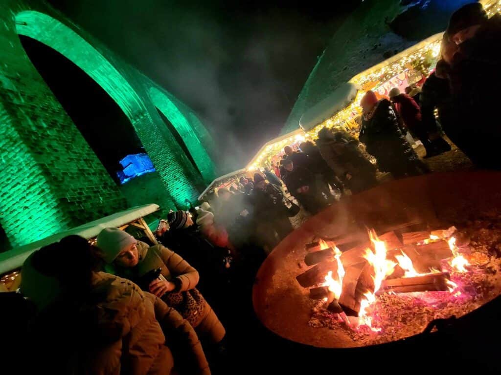 A tall arched stone viaduct illuminated in green with a large raised fire pit with a fire and people standing around with hats and heavy coats holding mugs