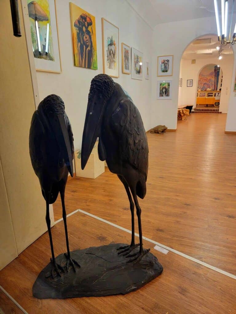 An art gallery with paintings, and a sculpture of two maribou storks in the window