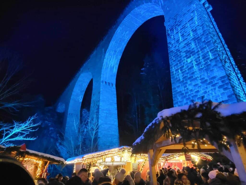 A tall arched stone viaduct illuminated in blue with several Christmas market stalls with snowy roofs, and a crowd of people