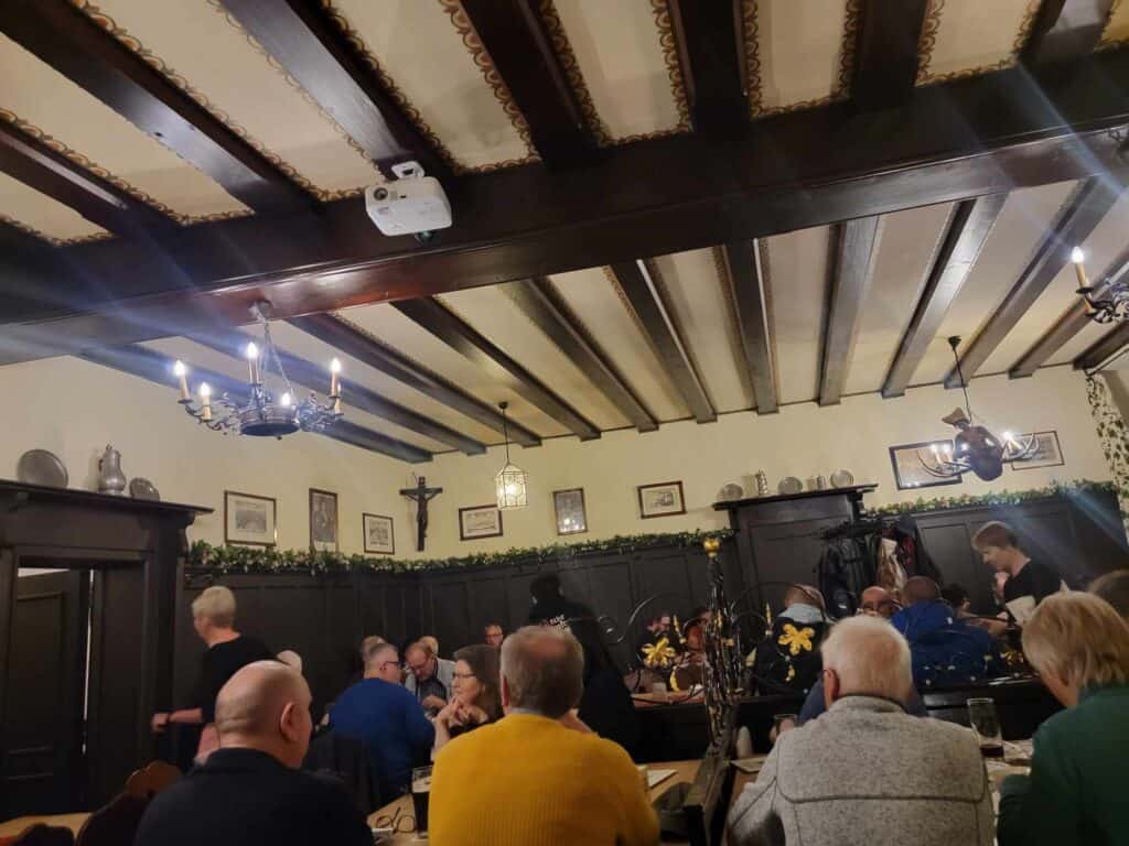 A room with tables of people eating and drinking and beamed ceiling