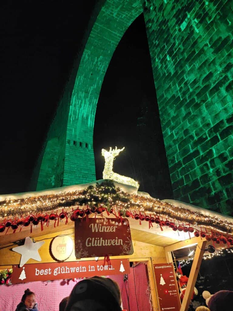 A tall arched stone viaduct illuminated in green with a market stall that has a sign saying Winzer Glühwein and "best glühwein in town" with an illuminated white reindeer on the roof