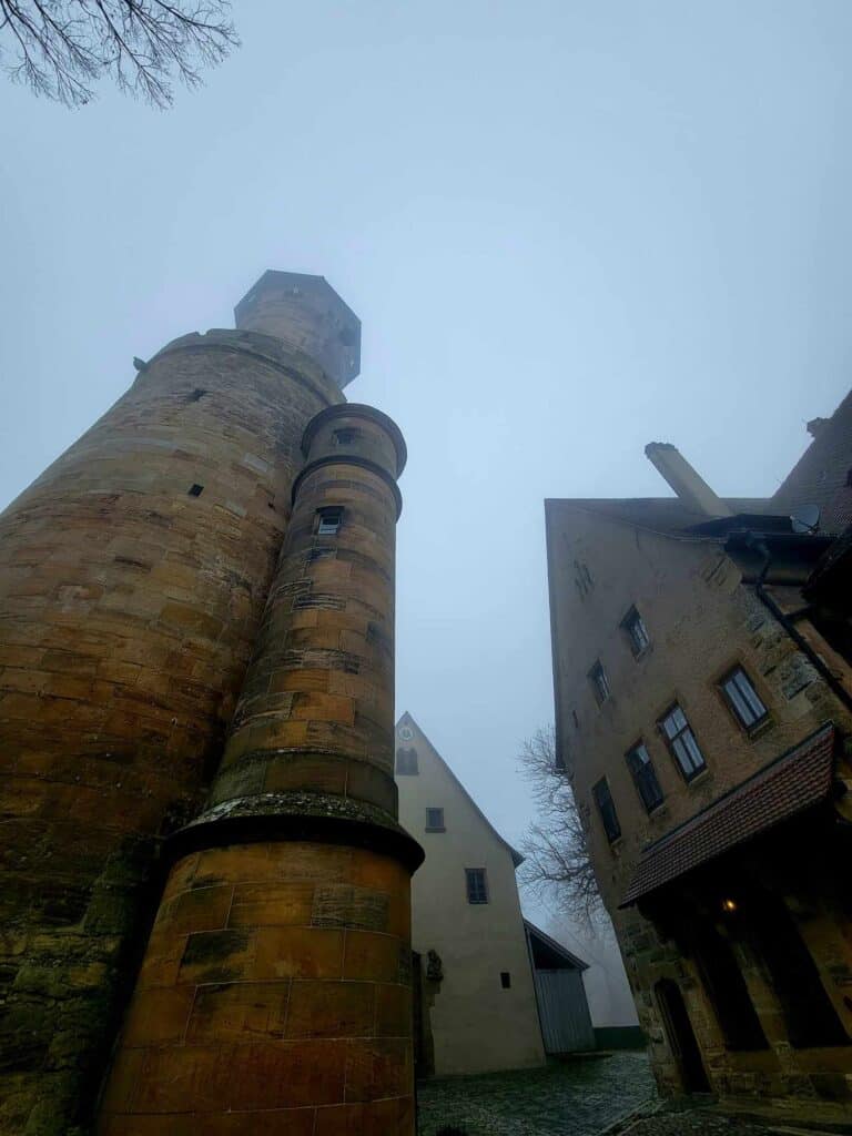 Two tall round towers side by side and a cobblestone path to the right with other peak-roofed stone buildings