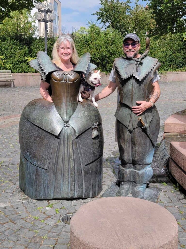 A couple standing behind bronze likenesses of medieval clothing. The woman holds a french bulldog