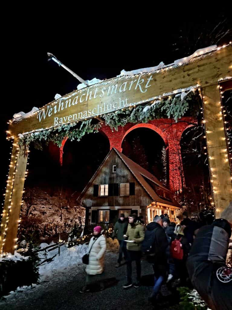 A square wooden arch that says Weihnachtsmarkt Ravennaschlucht with a chalet behind, and a tall stone viaduct illuminated in red