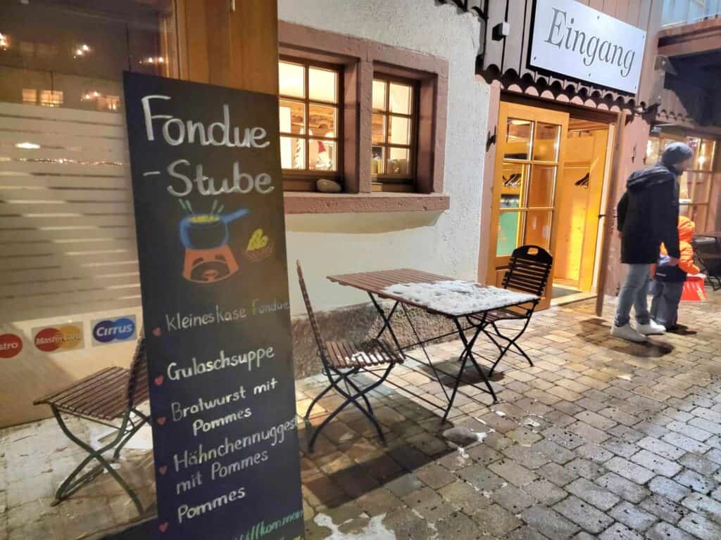 A small table and chairs covered with snow outside a restaurant with paned windows and a large blackboard sign that says "Fondue Stube" and a list of food like Gulaschsuppe, Bratwurst mit pommes, and Hahnchennugges mit pommes