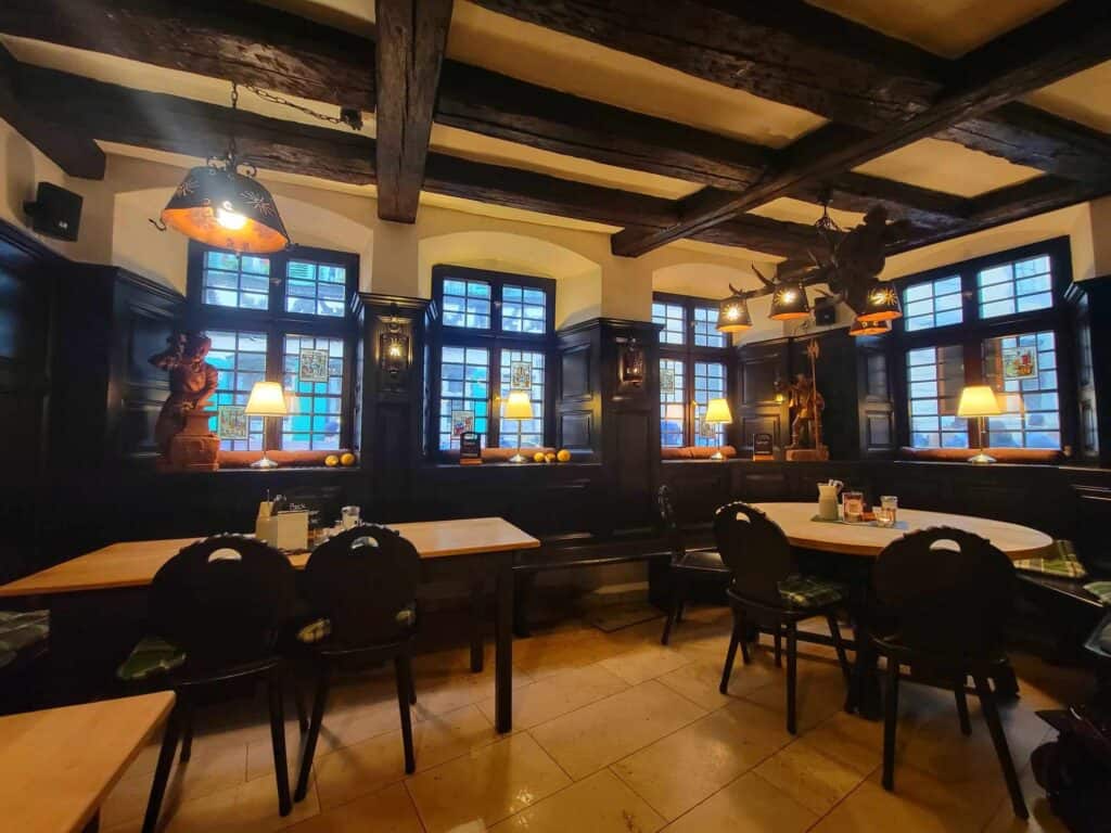 Super cozy tavern interior with dark beamed ceiling, paned windows with some stained glass, stone tiled floors and tables with wooden chairs