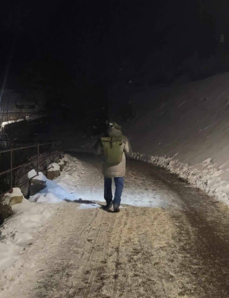 A man walking away from the camera on a snowy road. He has hiking shoes, jeans, a warm grey down coat, a hat and an olive green backpack