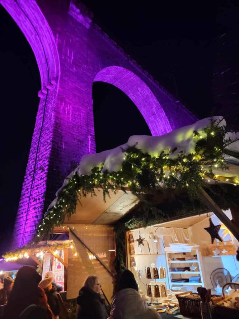 A tall arched stone viaduct illuminated in purple with a christmas stall decorated in greenery and white lights with snow on top.