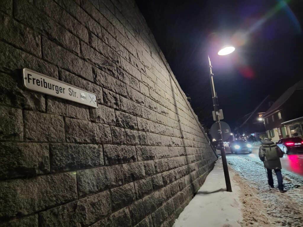 A high stone wall with a sign saying Freiburger Str. A man with a backpack walks on a snowy sidewalk by the road