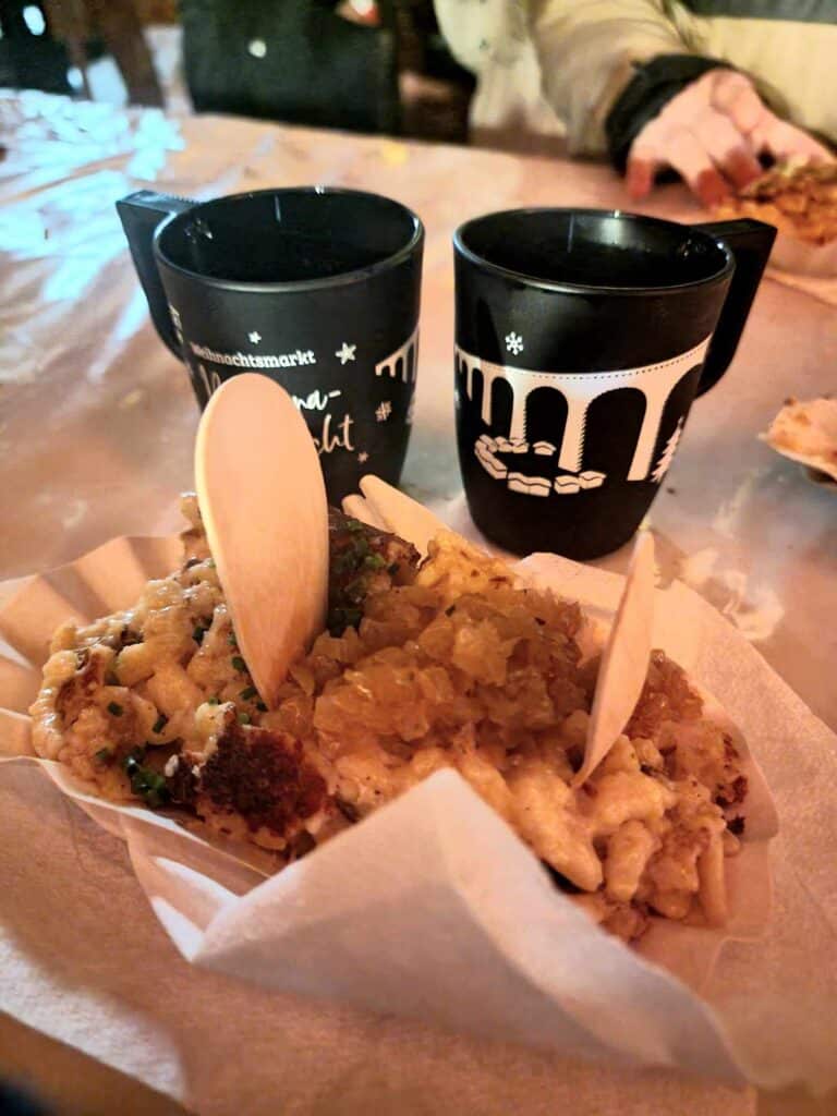 A paper plate piled high with cheese spaetzle and 2 wooden spoons. Two black mugs with an image of the viaduct, full of glühwein