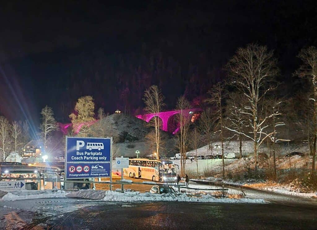 A blue parking sign that says Bus Parkplaz Coach Parking only and a parking lot with a bus full of people. A tall arched stone viaduct illuminated in pink in the background.