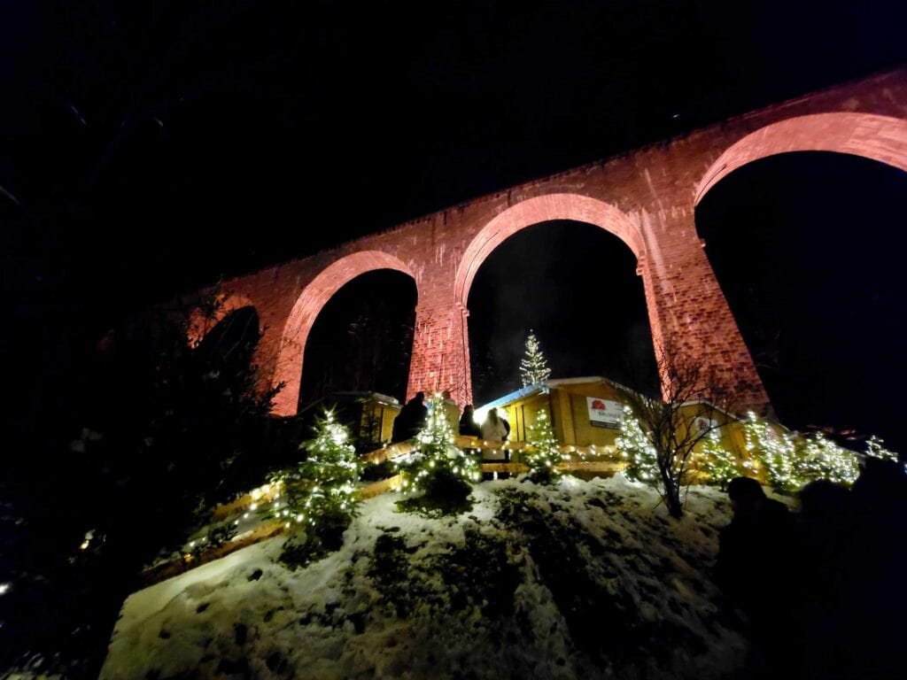 A tall arched stone viaduct illuminated in red with several evergreens covered in white lights and a wooden hut