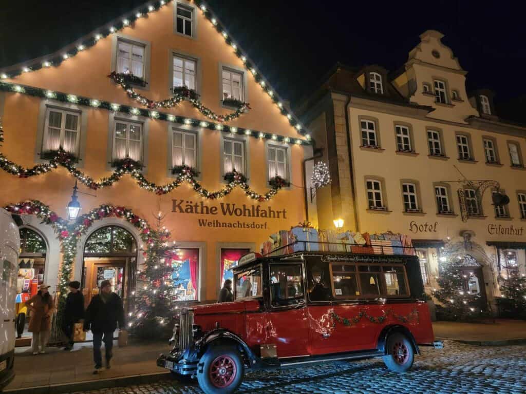 An old fashioned red car with a roof rack filled with wrapped presents in front of the Kathe Wohlfahrt Christmas village store covered in festive garlands