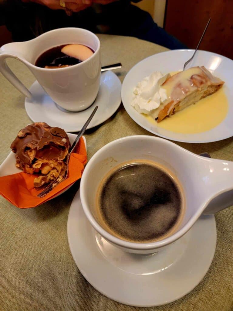 A cup of gluhwein with sliced apples in it, a plate of apple streudel with whipped cream and vanillasauce, a cup of coffee, and a chocolate schneeball