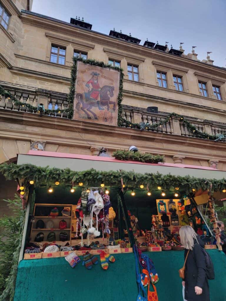 A Christmas market stall with knit items decorated in greenery in front of the town hall with a large image of a rider in a red coat on a brown horse up on two legs