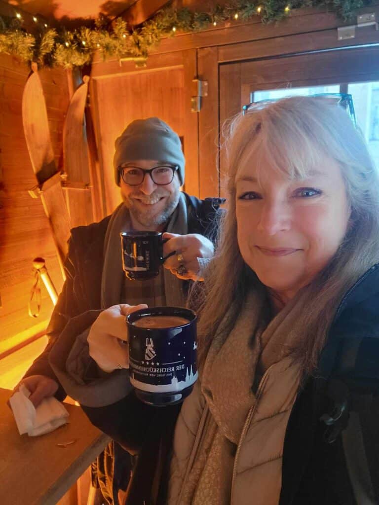 A smiling couple holding mugs of gluhwein in a cozy looking enclosure with lit garland