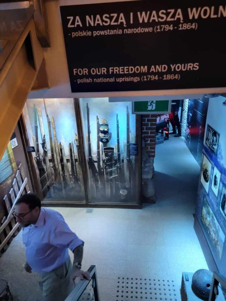 A man descends a staircase into a museum showcasing glass cabinets of rifles and war memorabilia and photographs. A sign reads FOR OUR FREEDOM AND YOURS polish national uprisings 1794-1864