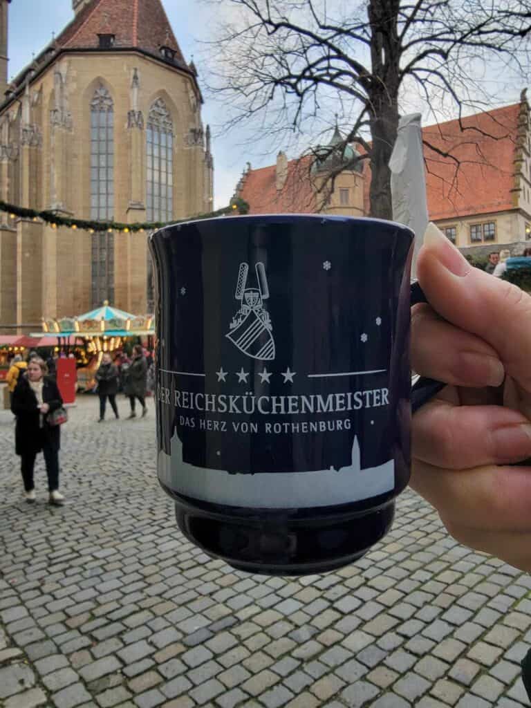 A woman's hand holding a dark blue mug in front of a medieval church