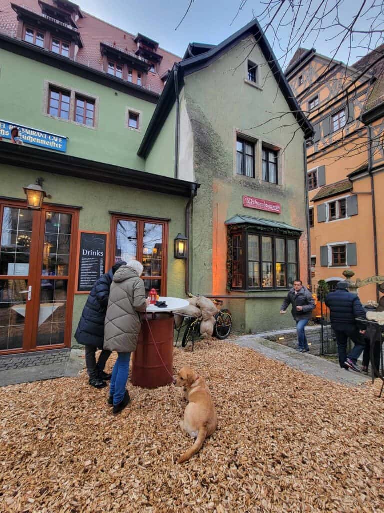 Two people drinking gluhwein at an outside round table in front of a green medieval building, with a golden retriever laying at their feet