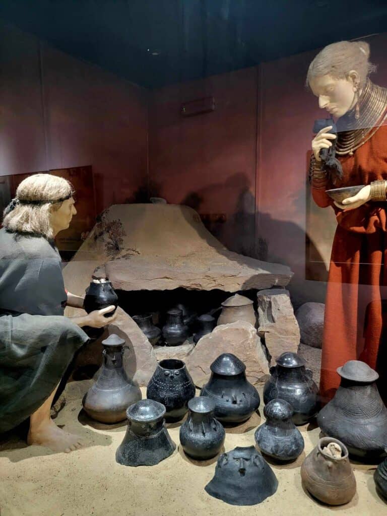 Museum figures of a man and a woman in tunics get ready to entomb a small basalt funerary vase in a large earthenware enclosure