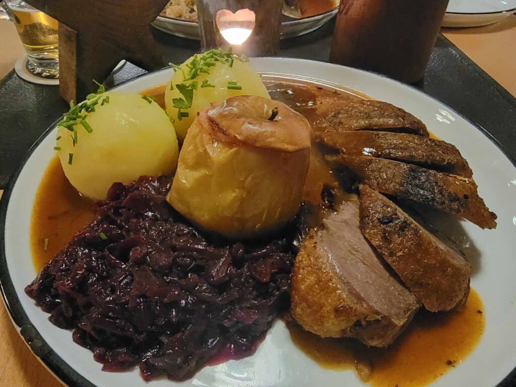 A plate of goose breast with gravy, red cabbage, potato dumplings and a baked apple on a white plate 