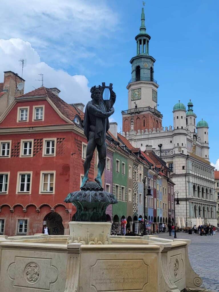 A white stone fountain base with a bronze statue of a man with a laurel wreath playing a lute. Behind is a colorful series of Renaissance row houses and an elaborate towered Town Hall.