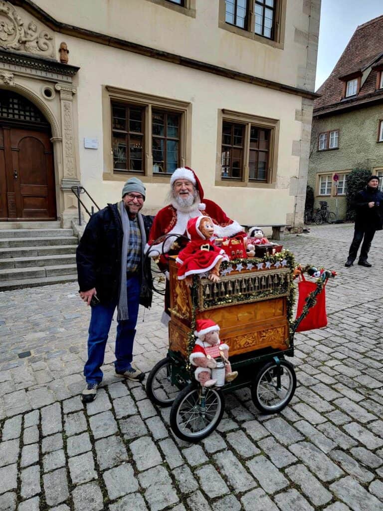 A man smiles standing next to Santa Claus with a stuffed santa monkey and an organ cart