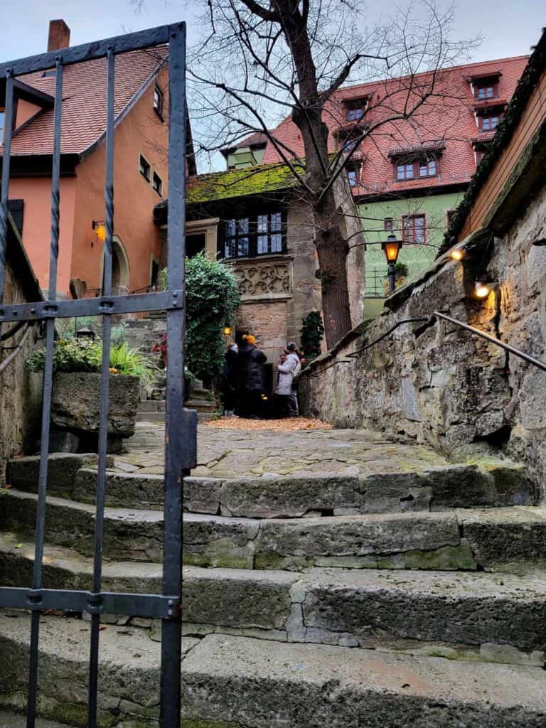 Stone stairs lead through an iron gate with quaint colorful houses and a stone wall backdrop. Peple are inside around a table