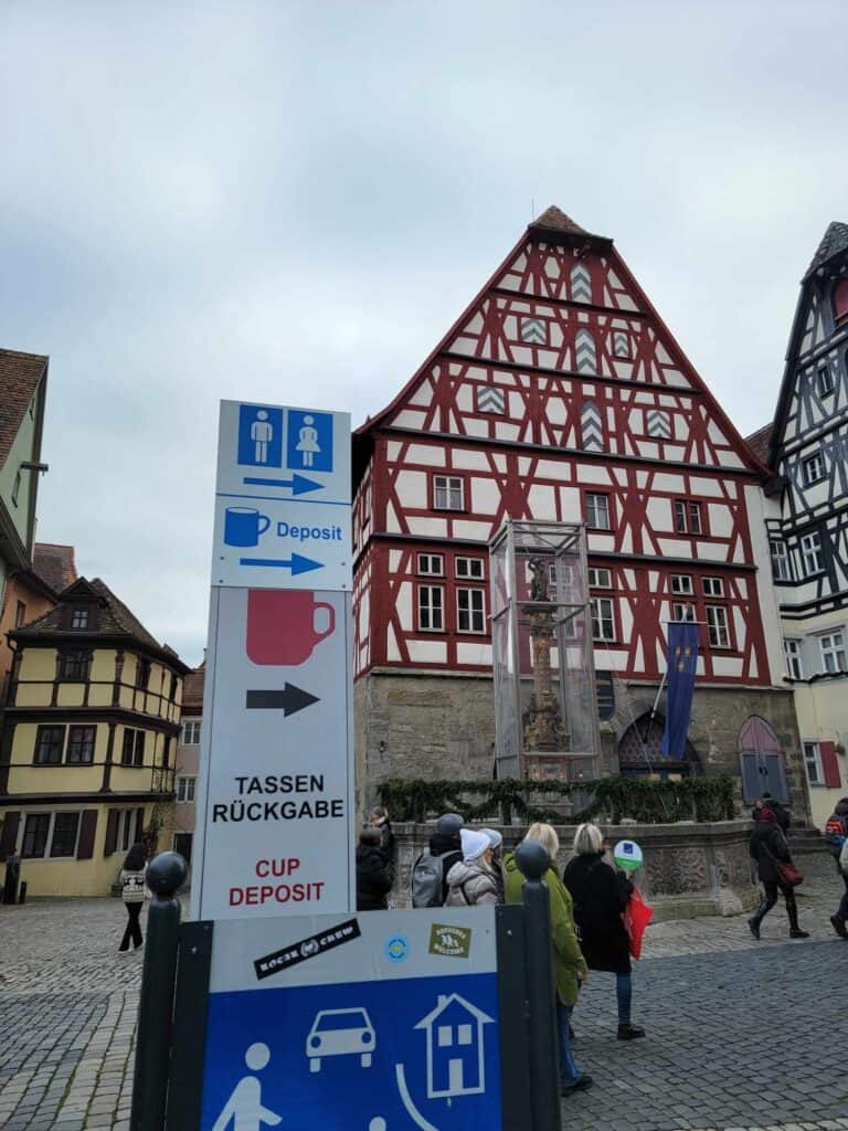 A tall medieval house half timbered in red and white in the background and a sign that says cup deposit with an arrow and a picture of a cup