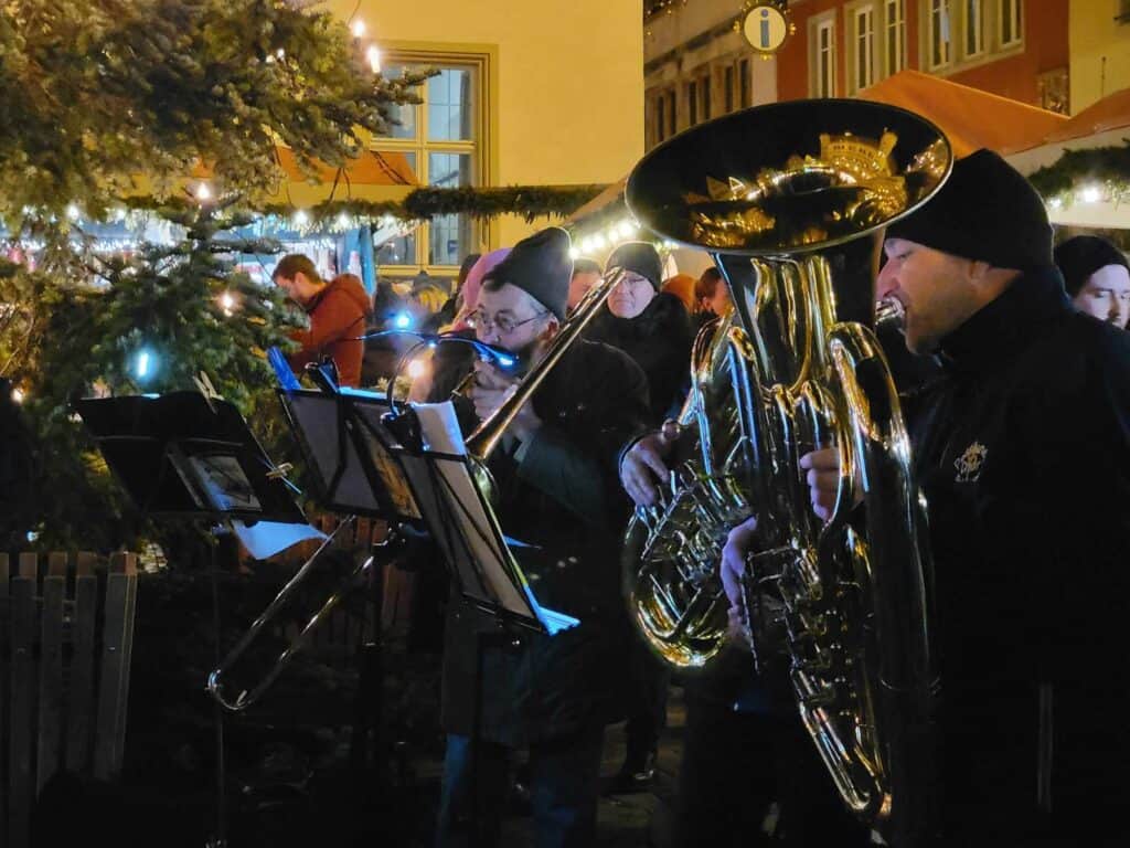 A trombone player and a tuba player outside in front of a lit christmas tree and onlookers