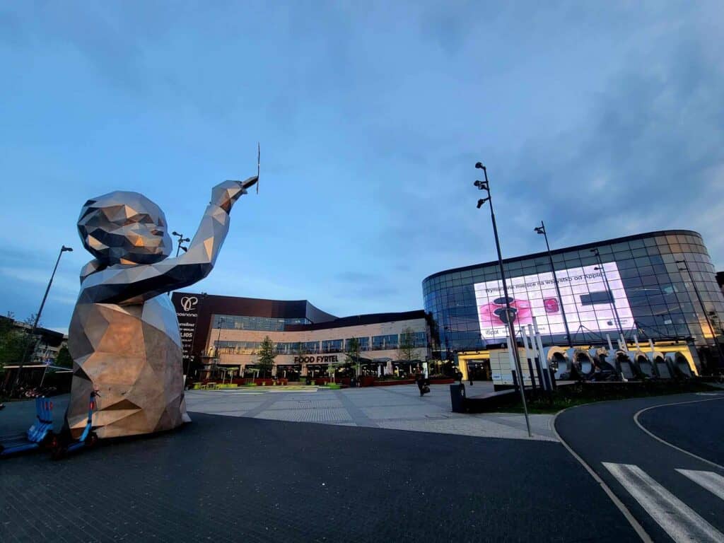 The outside of a modern shopping mall with metal letters in the foreground spelling posnania. An angular sculpture of a little girl who appears to draw a line in the sky with a pencil