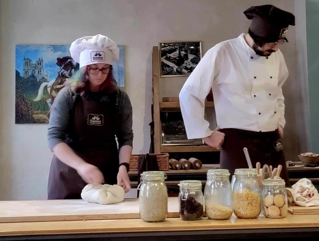 A woman in a white chef's hat pounds a ball of dough on a large wooden board. Jars and vessels full of ingredients are in the fore, and a man in a white coat and black chef's hat looks at them.