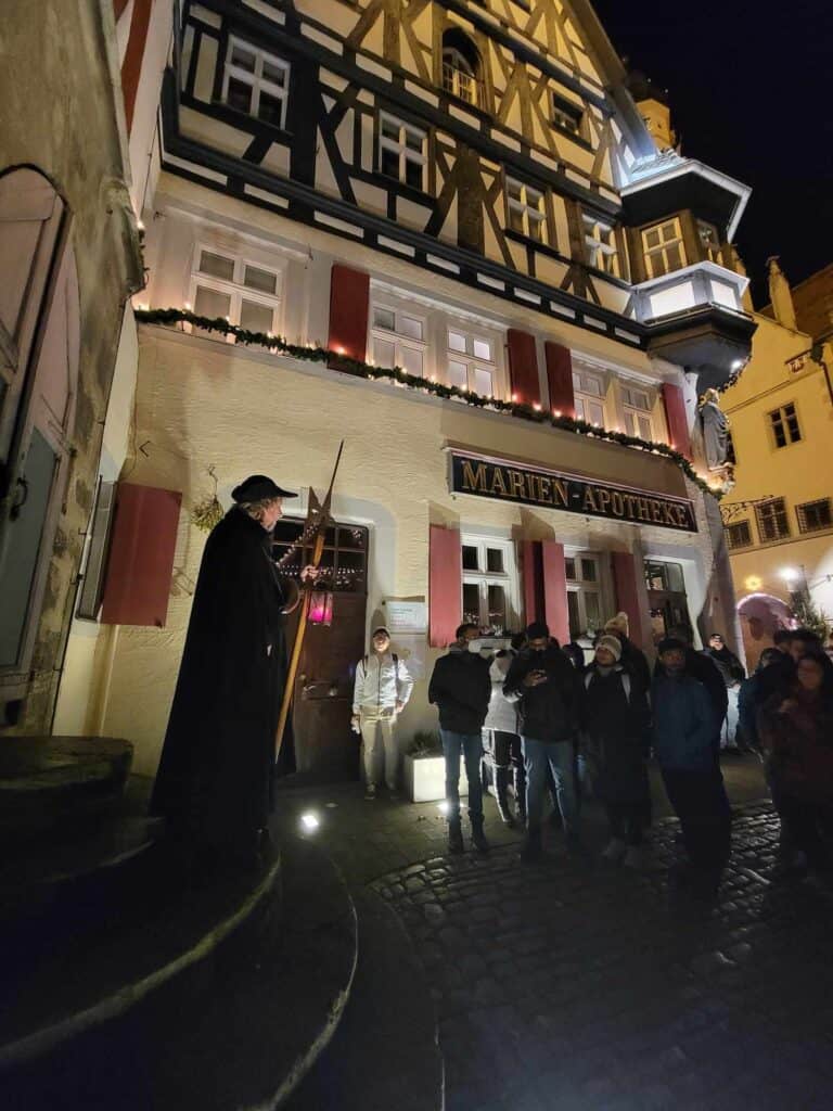 The night watchman in a black cloak and hat holding a halberd and a glowing lantern with a group of people looking on 