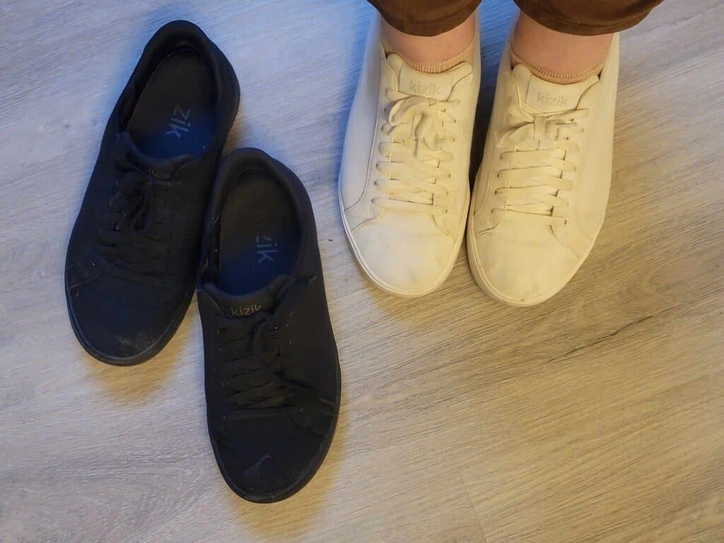 A woman's feet inside a pair of white sneakers with an empty pair of black sneakers next to her