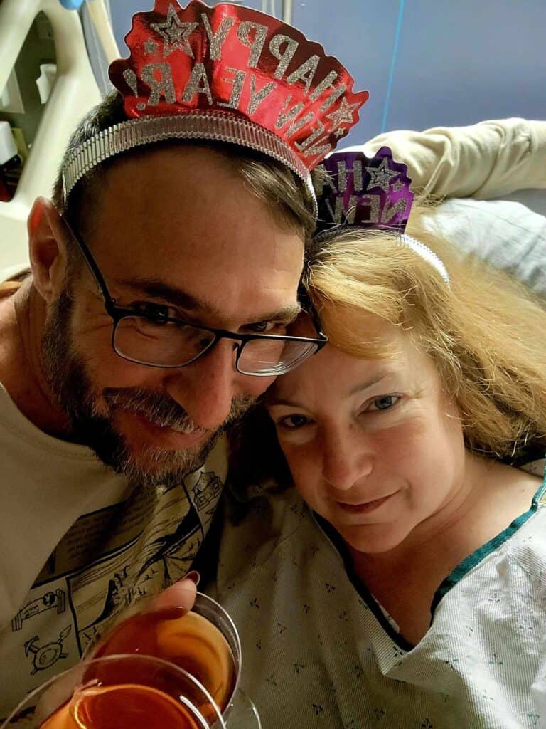 A woman in a hospital bed with a man next to her. They are holding glasses of sparkling apple juice and wearing headbands that say happy new year