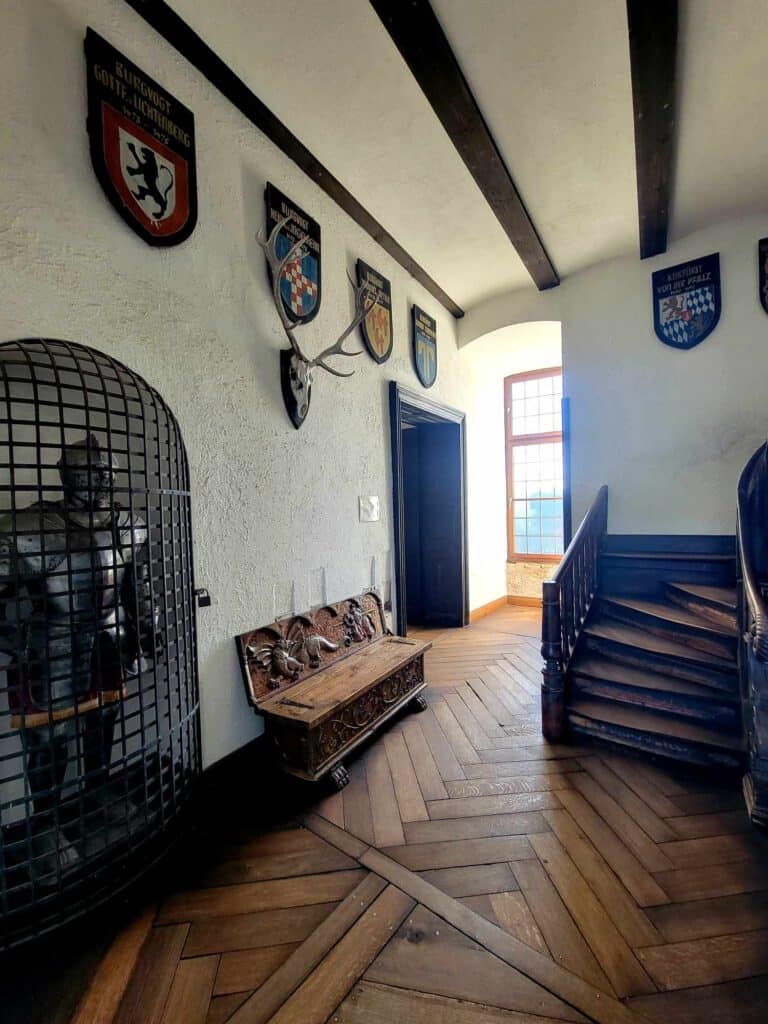 A white plastered room with exposed beams, coats of arms decorate the walls, along with a deer skull and antlers. There is a suit of armor in a closet-like cage, herringbone pattern wood floors and a wooden staircase.