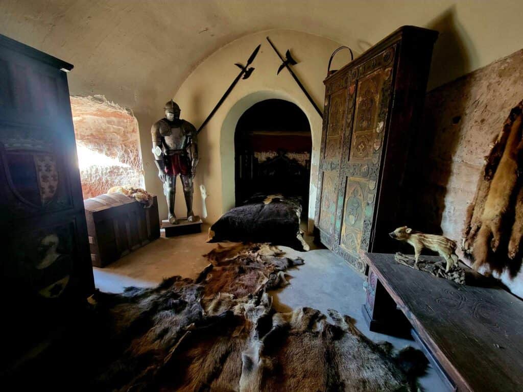 A bedroom with a suit of armor and two halberds over the bed which has an animal skin covering it. Animal skin rugs on the floor and an ornate carved wooden wardrobe.