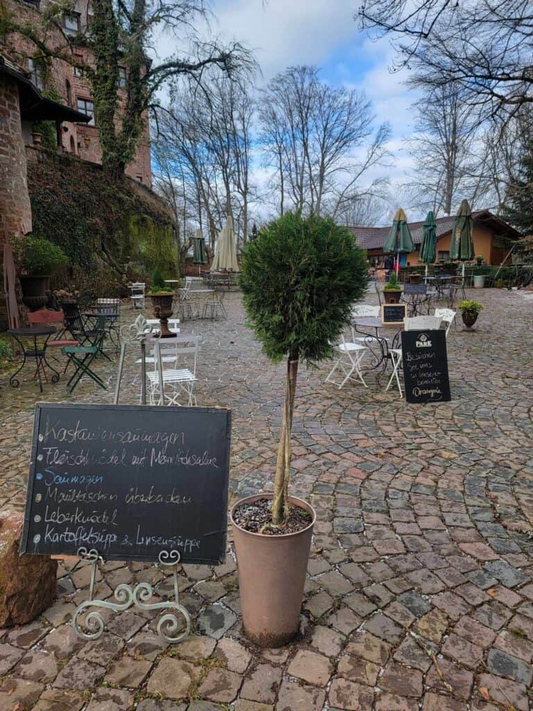 A cobblestone terrace with white cafe tables and chairs next to the castle. There are bare trees and closed umbrellas. Chalkboard sign lists food available in German