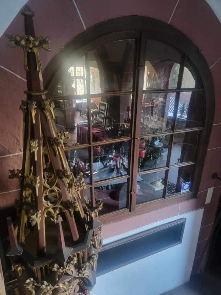 Looking past an ornate wooden carving of a spire and through a glass window into a dining room with red table coverings