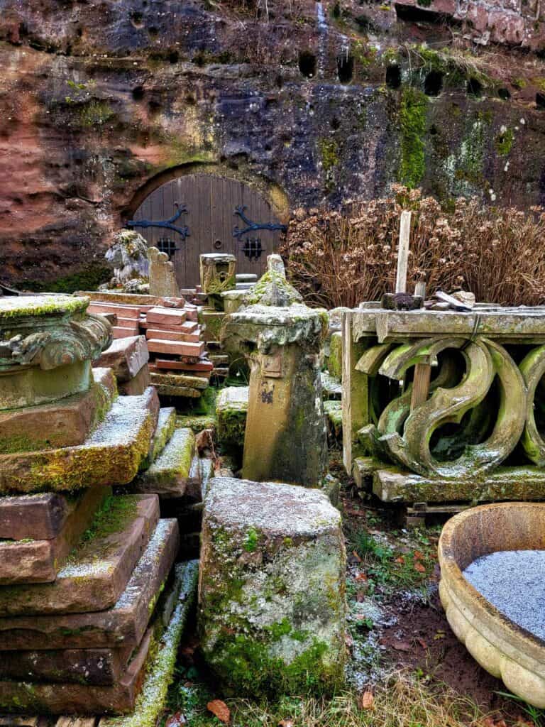 A big pile of architectural elements tiles and traceries, and a lion statue all covered in lichen and moss sitting in front of a stone wall with an arched dark wood door