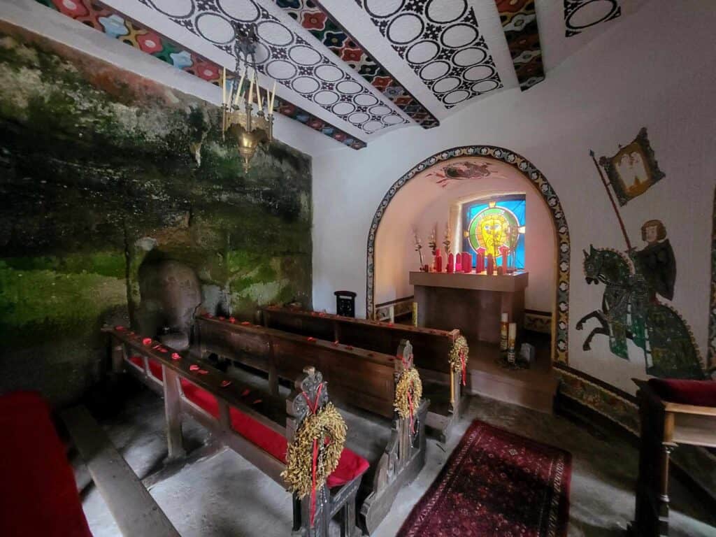 A small chapel with frescoed walls and ceiling and three pews with a small altar covered in red candles and a piece of blue and yellow stained glass