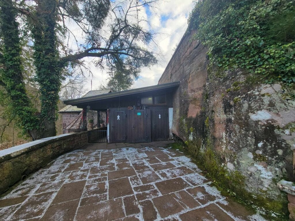 A stone terrace next to a high stone wall. There are two restrooms in a small wooden shed at the end of the terrace