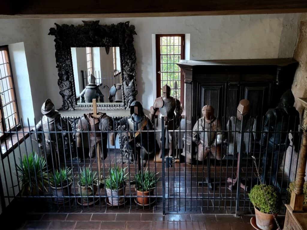 A line of suits of armor behind a short wrought iron railing. There is a large ornate mirror, a leaded glass window, and some houseplants