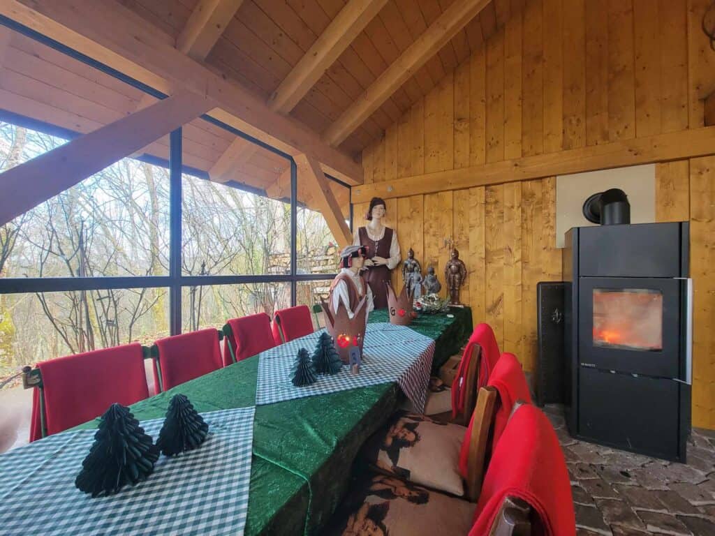 A long table with a green checkered tablecloth and red candles with wooden chairs draped in red blankets. Two mannequins are dressed in medieval clothing and stand by the window.