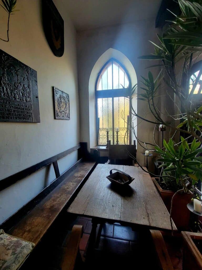 A narrow room with white walls and an arched gothic window. There is a long wooden bench and a rustic wooden table and chair.