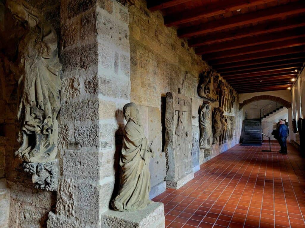 A long corridor with red tile floor and stone statues representing religious figures lining the stone wall on the left