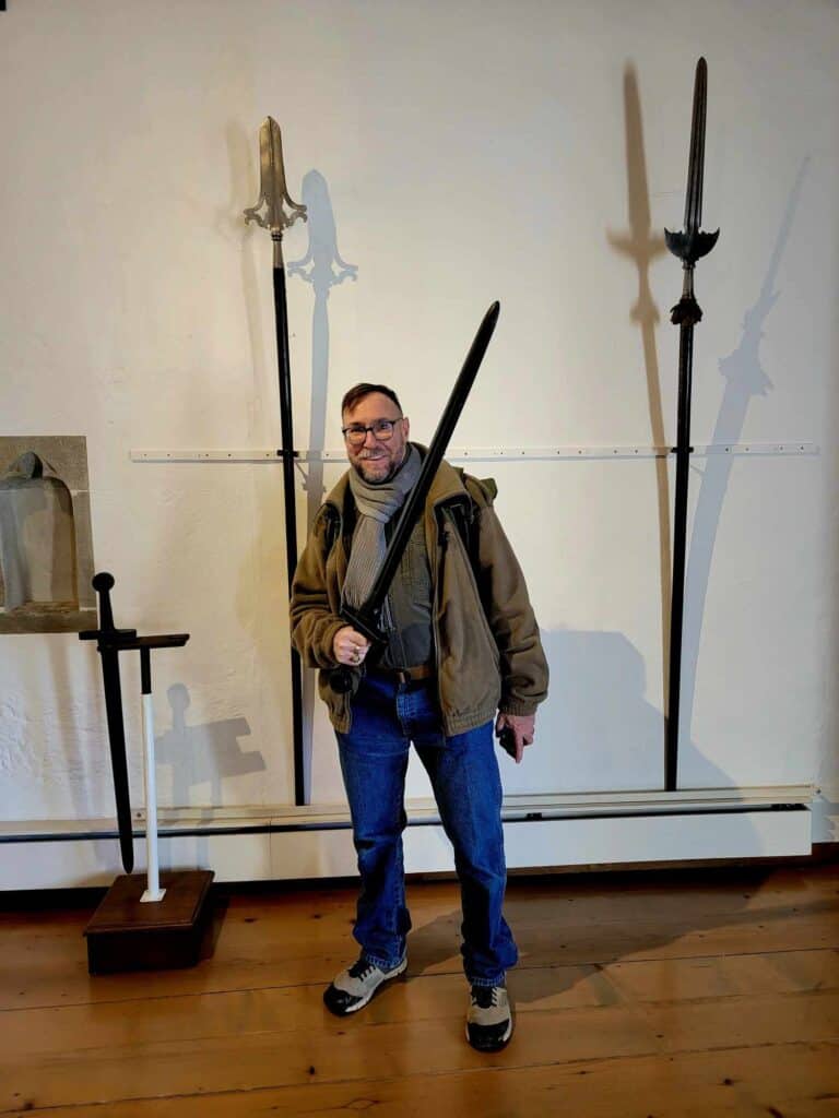 A man smiling and holding a medieval sword replica with two halberds on the wall behind