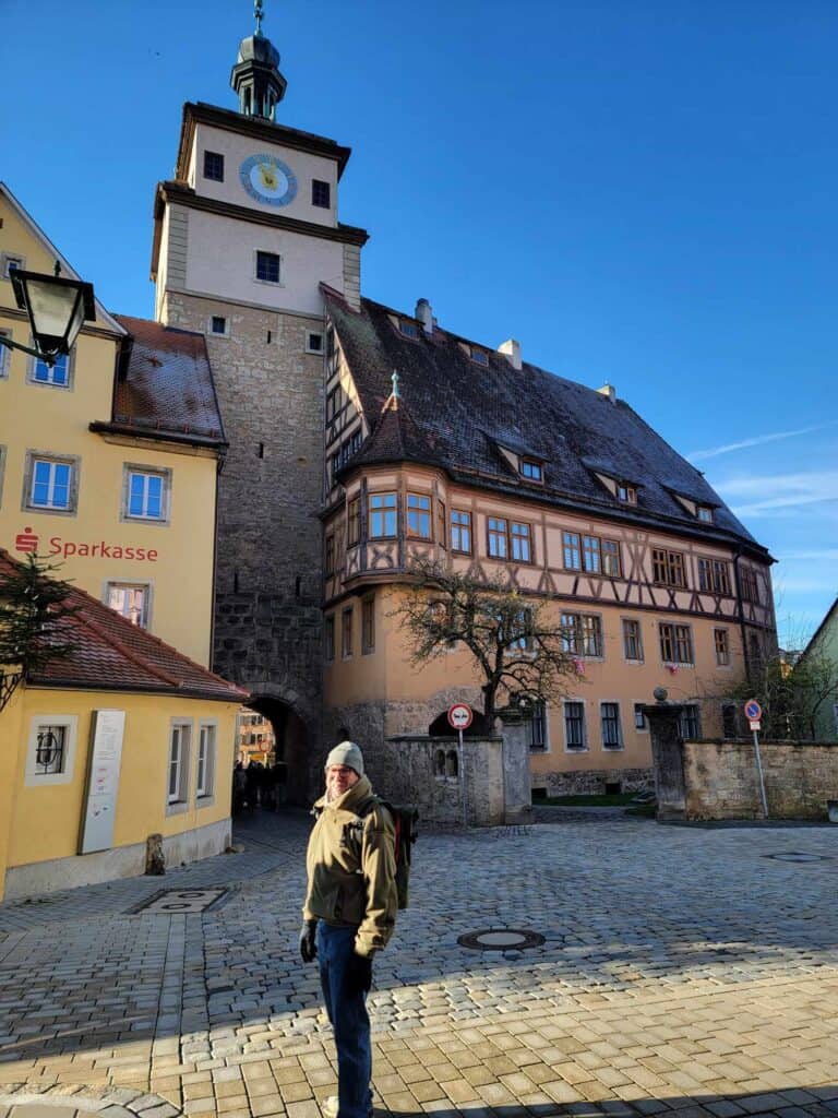 A man stands on a cobblestone square with a timbered house and tower with clock behind