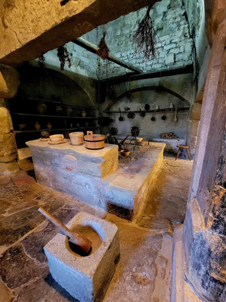 A medieval kitchen with stone island covered with pots, an arched stone niche with pots hanging, and herbs hanging from the ceiling