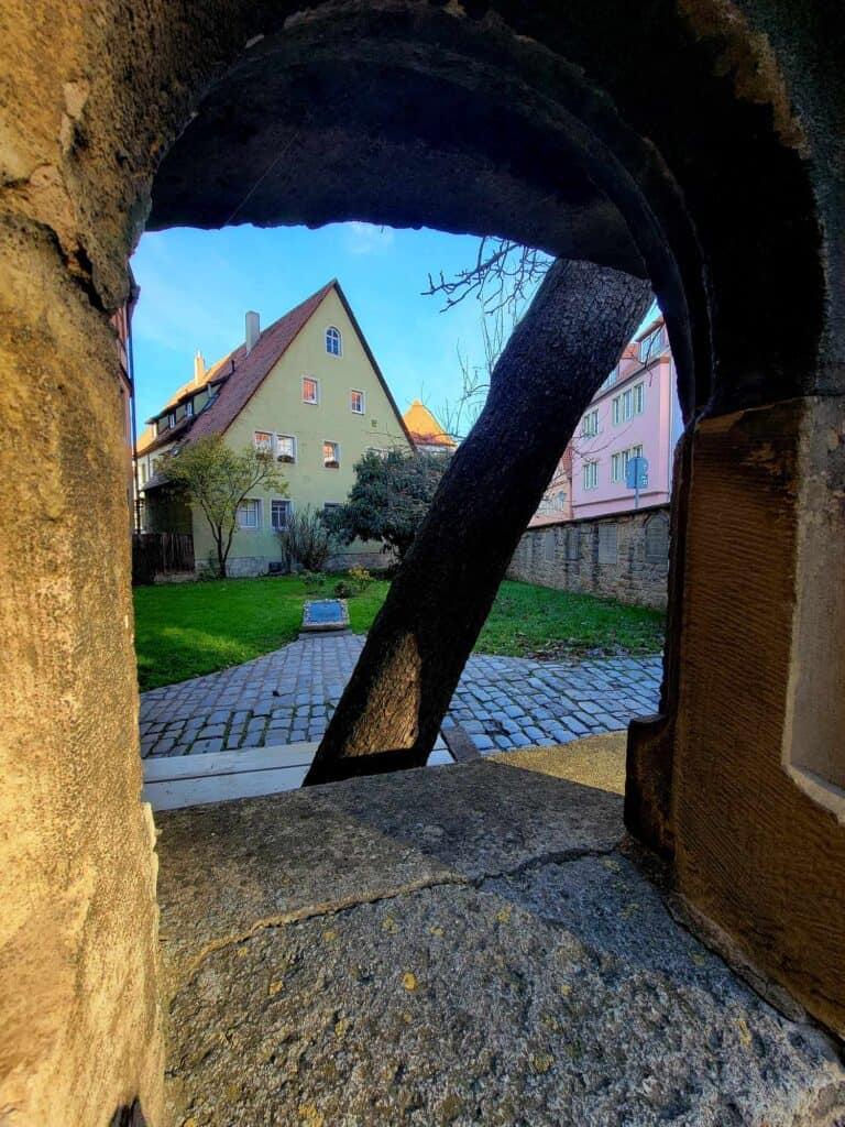 Looking through a small window in a stone wall at a cobblestone path, a green grass walled garden